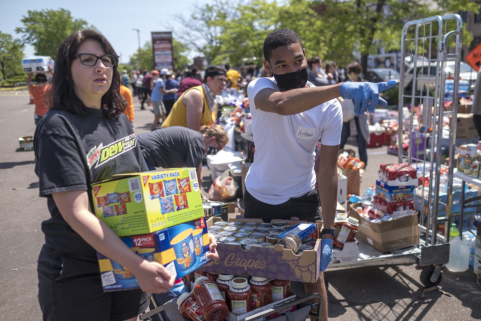 A group of people at a food distribution event with tables and carts filled with boxes, cans, and jars. One person holds a box of "Variety Pack" chips while another points off-camera. Trees and more people are visible in the background
